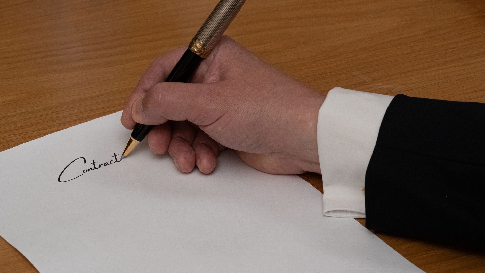 Main d’une personne en costume écrivant le mot « Contract » au stylo sur une feuille blanche posée sur une table en bois.