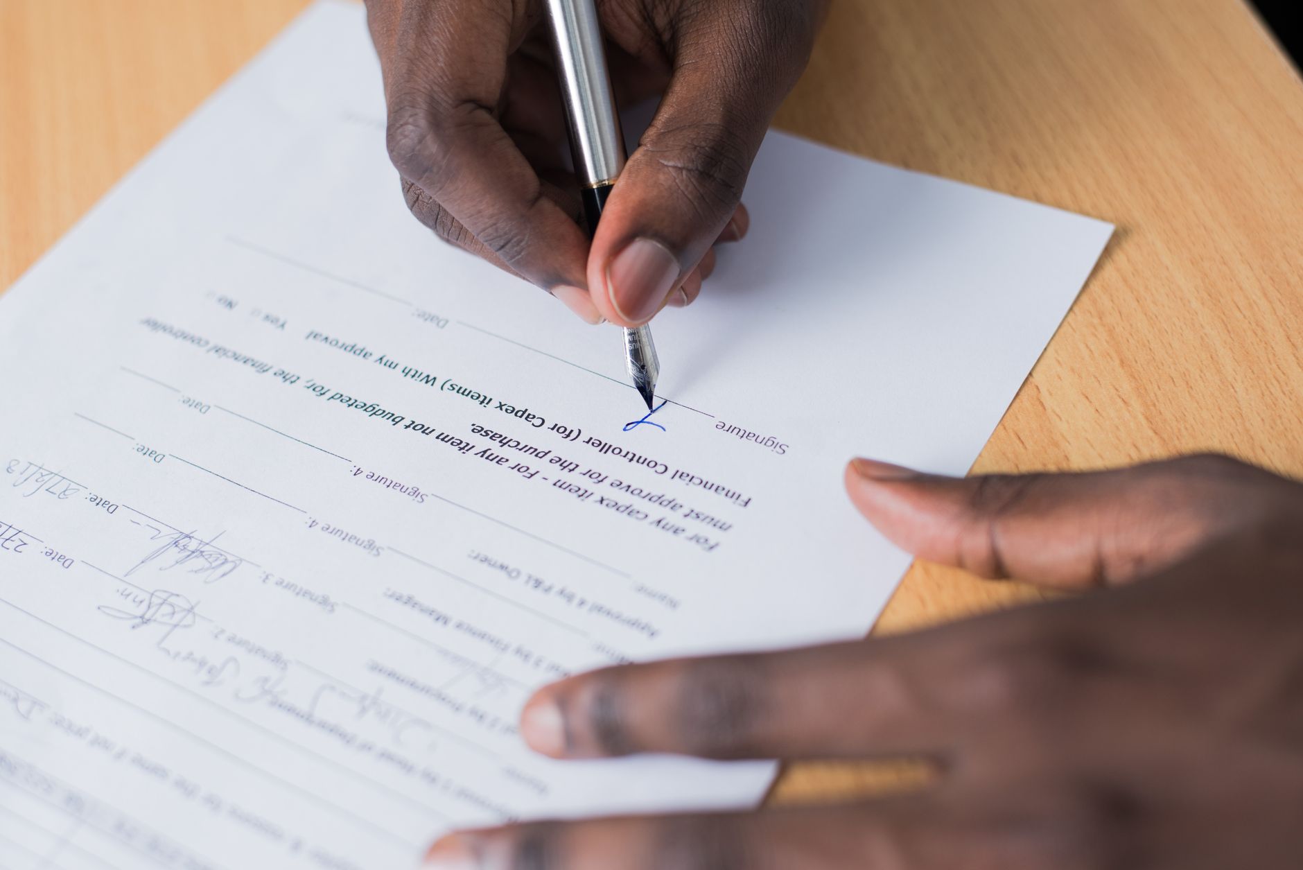 Une main signe avec un stylo-plume un formulaire à lignes de signature posé sur un bureau en bois, l’autre main tenant la feuille.